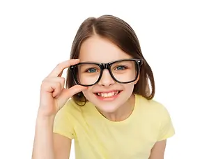 A young girl with brown hair is smiling and holding a pair of black glasses up to her face.