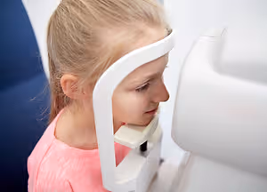 A girl in a pink shirt looking into an eye exam device in a room