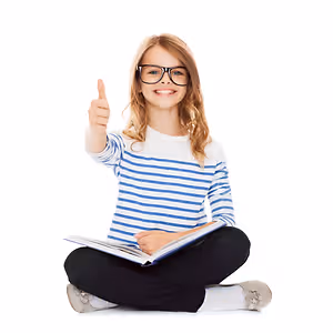 A girl wearing glasses and a striped shirt is sitting on the floor and smiling while giving a thumbs up with her left hand and holding a book with her right hand.