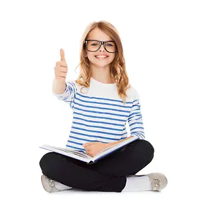 A girl wearing glasses and a striped shirt is sitting on the floor and smiling while giving a thumbs up with her left hand and holding a book with her right hand.