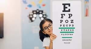 A young girl with glasses is posing for a picture behind an eye chart in a colorful room.