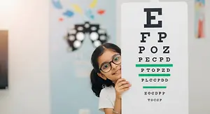 A young girl with glasses is posing for a picture behind an eye chart in a colorful room.