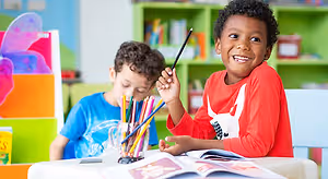 Two children sitting at a desk with one holding a pencil and smiling, and the other holding a cup with colorful pencils. There is a book and other objects on the desk in front of them, and a bookshelf with books and other items in the background.