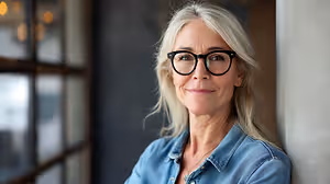 Portrait of a mature woman wearing glasses and a denim shirt, smiling and looking out of a window with a bright, warm light.