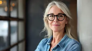 Portrait of a mature woman wearing glasses and a denim shirt, smiling and looking out of a window with a bright, warm light.