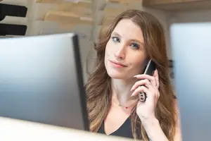 A woman wearing a necklace is talking on a cell phone while sitting in front of a computer monitor.