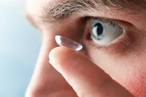 A close up of a man wearing glasses holding a contact lens in his finger