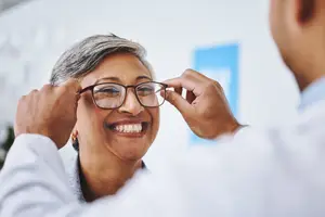 An elderly woman is wearing eyeglasses and smiling while looking at a doctor