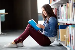 A woman reading a book on the floor near a shelf of books in a library