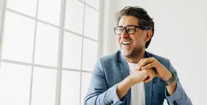 A smiling man in a suit with glasses on and his hands clasped in front of him