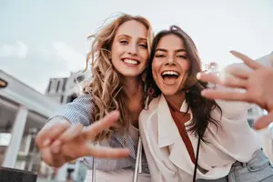 Two women are standing next to each other and smiling while taking a selfie with their cellphones.