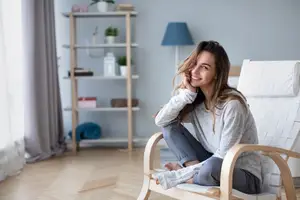 A woman is sitting in a chair and smiling in a room with a shelf and a lamp.