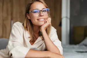 A smiling woman with glasses and a white shirt is posing for a photo while lying on a bed.