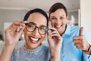 A woman is adjusting her glasses and smiling while another woman is giving her a thumbs up