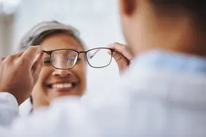 An older woman trying on a pair of glasses in front of a smiling man in a white coat