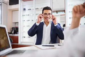 A man wearing a blue blazer and white shirt is adjusting his glasses in an office room with a desk, laptop, and mirror.
