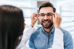 A man is trying on a pair of glasses with the assistance of a woman in a white coat.