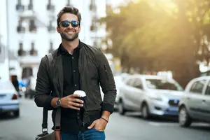 A man in a jacket is walking down a street, smiling and holding a cup of coffee. He is also wearing sunglasses, a watch, and a bracelet. Behind him, there are several cars parked on the side of the road, and a building with many windows is in the background. The sun is shining brightly, and the light is reflecting off the cars and the building.