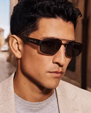 A close-up view of a man wearing sunglasses and a gray shirt with a blazer while looking to the side in a street setting.