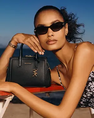A woman wearing sunglasses and a floral dress holds a black leather handbag by the handle.