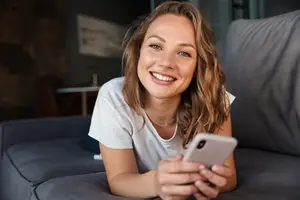 Young woman sitting on a couch, holding a cell phone, and smiling