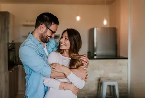 A man in a blue shirt and a woman in a white shirt are hugging each other in the kitchen.