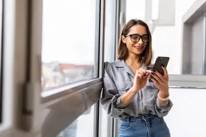A smiling woman with glasses is standing in front of a window, holding a cell phone and looking at something. She seems to be in a room with a glass window that reflects her image, and she is wearing a bracelet on her wrist. Behind her, there is a blurry view of a building with a roof, and the sunlight is shining on the glass window.