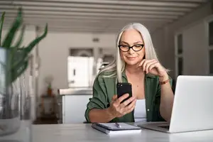 A woman with white hair wearing glasses and a ring is smiling while looking at her cell phone in front of a desk with a laptop and a plant in a vase.