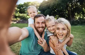 A family is taking a selfie on a sunny day in a park with trees in the background.