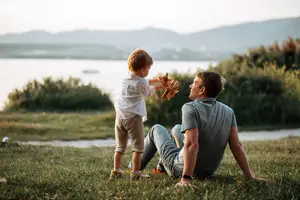 A man sitting on a lawn is looking at a toddler standing in front of him. The toddler is holding a wooden toy and seems to be playing with the man. Behind them, there is a lake with a boat sailing on it. In the distance, there are mountains, trees, and plants.