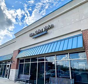 Blue Valley Vision is the name of a store with a blue awning over the entrance, a bench on either side, and a clear view of cars parked outside in a parking lot.