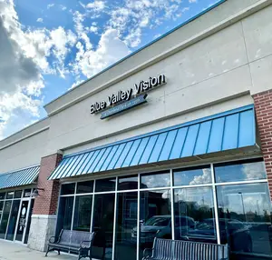 Blue Valley Vision is the name of a store with a blue awning over the entrance, a bench on either side, and a clear view of cars parked outside in a parking lot.