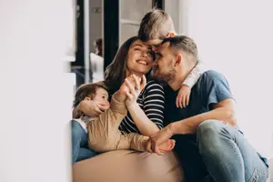 A happy family of four sitting in a cozy living room, with the father and mother smiling and looking at each other while the two children sit on their laps.