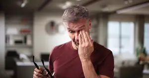 A man in a burgundy shirt is holding glasses and touching his face with his hand.