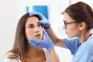 A woman with blue gloves examines a patient's eye in a medical setting.
