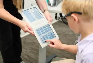 A young boy is sitting in a chair and interacting with a device held by a person wearing glasses and a bracelet. The device has a blue and white screen with numbers and letters, and the boy is pressing a button. The person is wearing black pants and a watch on their wrist, and the boy is wearing a white shirt and glasses.