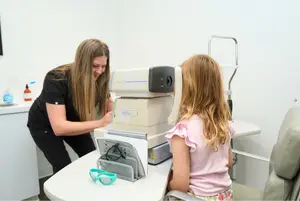 A woman helps a young girl use an eye exam machine at an optometrist's office.
