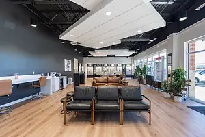 An empty waiting room in a medical clinic with wooden floors, black leather chairs, and potted plants on either side.