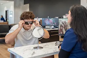 Man adjusting sunglasses while a woman helps him in a store with a monitor and mirrors in the background.