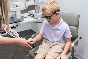 A boy wearing glasses is being examined by an optometrist while holding a tablet