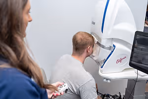 A man is undergoing an eye test while sitting and wearing glasses, while a woman holds a remote control and observes him.
