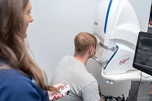 A man is undergoing an eye test while sitting and wearing glasses, while a woman holds a remote control and observes him.