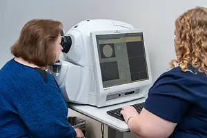 A woman is getting her eye examined with an autorefractor machine in a clinic while another woman is typing on a computer keyboard
