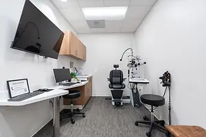 An empty examination room inside a building with a television, cabinets, a monitor, a keyboard, and a sign on the desk, a black chair, and a brown leather seat on the right corner.