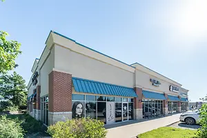 The front view of a building with a blue awning and a glass window that shows a reflection of a woman and some trees, and a white car parked on the side.