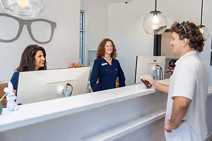 Three people are standing at the reception desk of an office with a man standing next to a woman with glasses and a woman smiling next to her with a bunny toy on the counter.