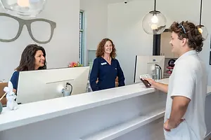 Three people are standing at the reception desk of an office with a man standing next to a woman with glasses and a woman smiling next to her with a bunny toy on the counter.