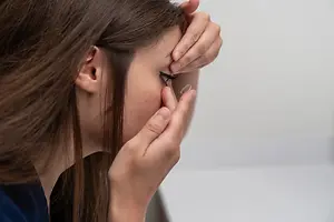 A woman is applying a contact lens to her eye while looking in a mirror