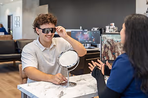 A man is trying on sunglasses and looking in a mirror while a woman looks at him in a medical office