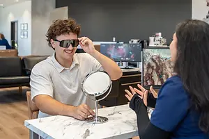 A man is trying on sunglasses and looking in a mirror while a woman looks at him in a medical office